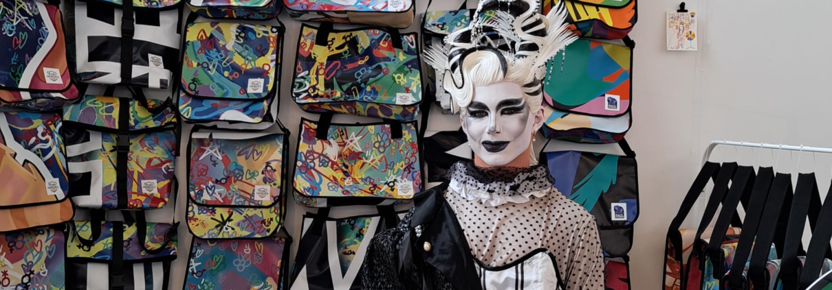 Person in schwarz-weißem Kostüm vor einer Wand mit bunten Upcycling-Taschen von PrideHEROES.
