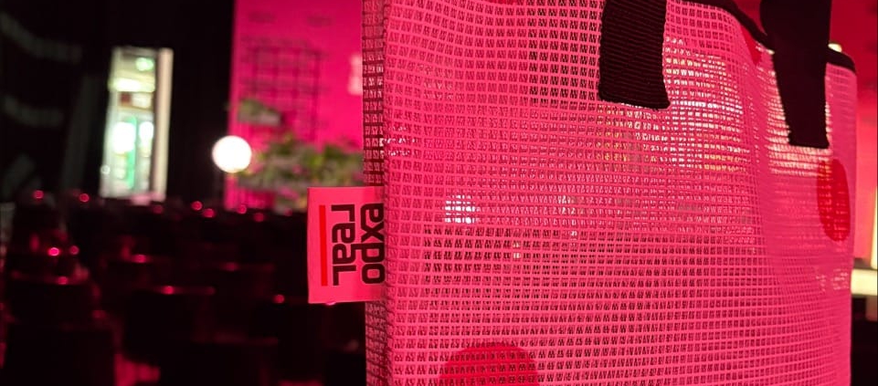 Einkaufstasche aus einem EXPO REAL-Mesh-Banner mit großen Punkten und schwarzen Henkeln in einer pink beleuchteten Veranstaltungshalle